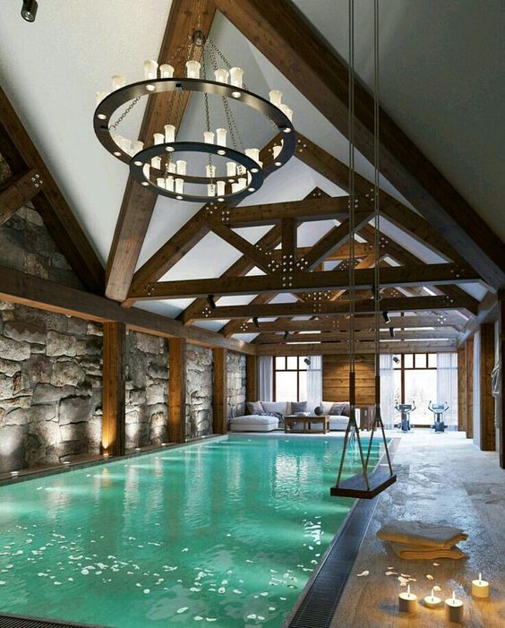 a fantastic pool house with an attic roof and wooden beams, a white sofa, a round chandelier, a large pool and a swing