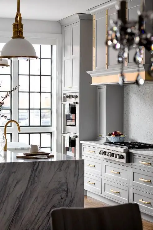 a glam grey kitchen with vintage cabinetry, gold handles and edging, a white marble kitchen island and brass fixtures