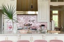 a glam tropical kitchen with white cabinetry, a pink tile backsplash and matching stools, pink tableware and pendant lamps