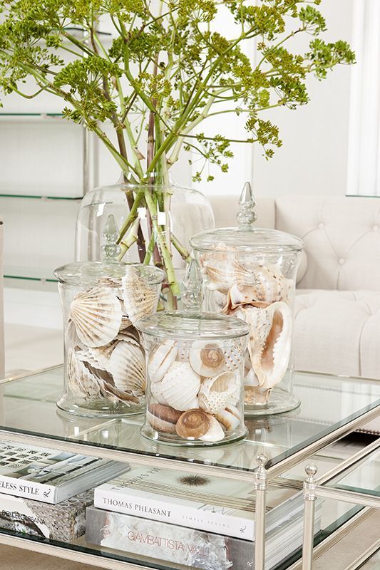 a glass coffee table with books, large jars with seashells and an oversized vase with some greenery