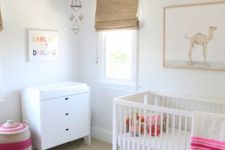 a light-filled nursery with white furniture including an IKEA Sundvik crib, a colorful rug, basket and bedding and wicker shades