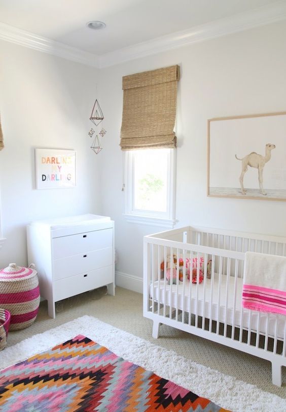 A light filled nursery with white furniture including an IKEA Sundvik crib, a colorful rug, basket and bedding and wicker shades
