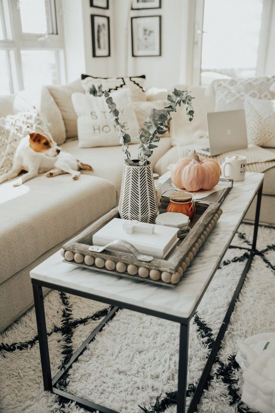 a marble coffee table, a wooden tray with beads, a vase with greenery, some candles and books and a pumpkin