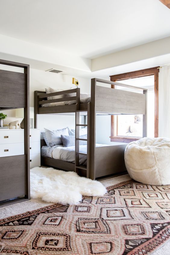 a modern boho kids' room with stained bunk beds, neutral printed bedding, layered rugs and a white pouf