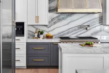 a modern glam kitchen with white and graphite grey cabinetry, a white stone backsplash and countertops, a glam hood and gold handles
