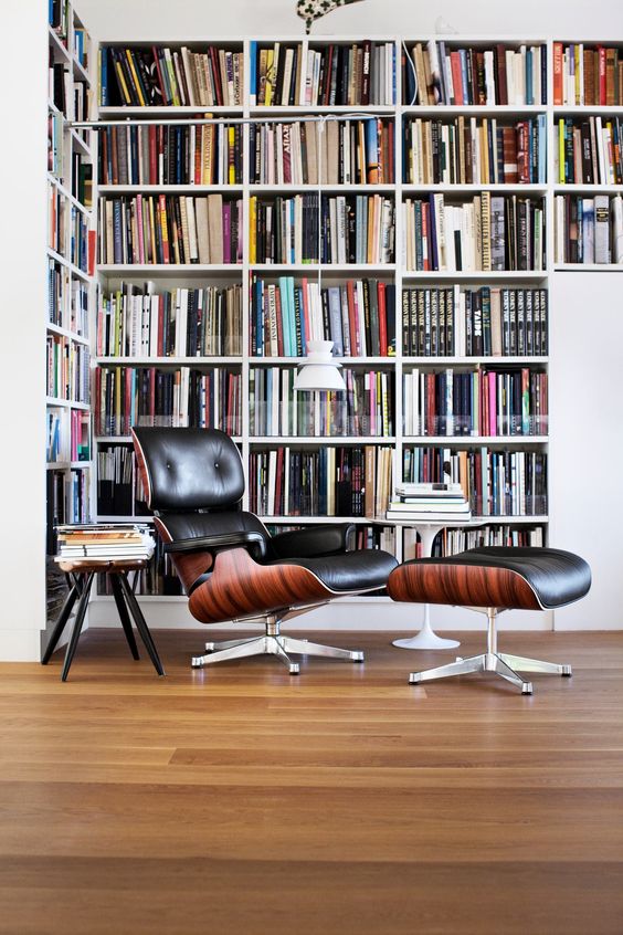 A modern home library nook with an oversized L shaped bookcase, a black leather chair with a footrest
