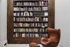 a modern home library with a bookcase in a wide wooden frame, with a comfy leather chair and a red pendant lamp