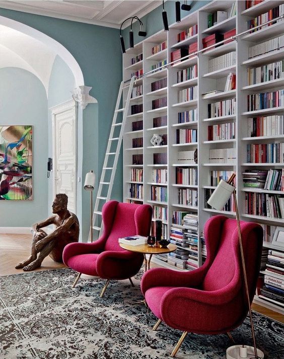a modern home library with an oversized white bookcase with steps and two chic chairs in red for a cozy nook