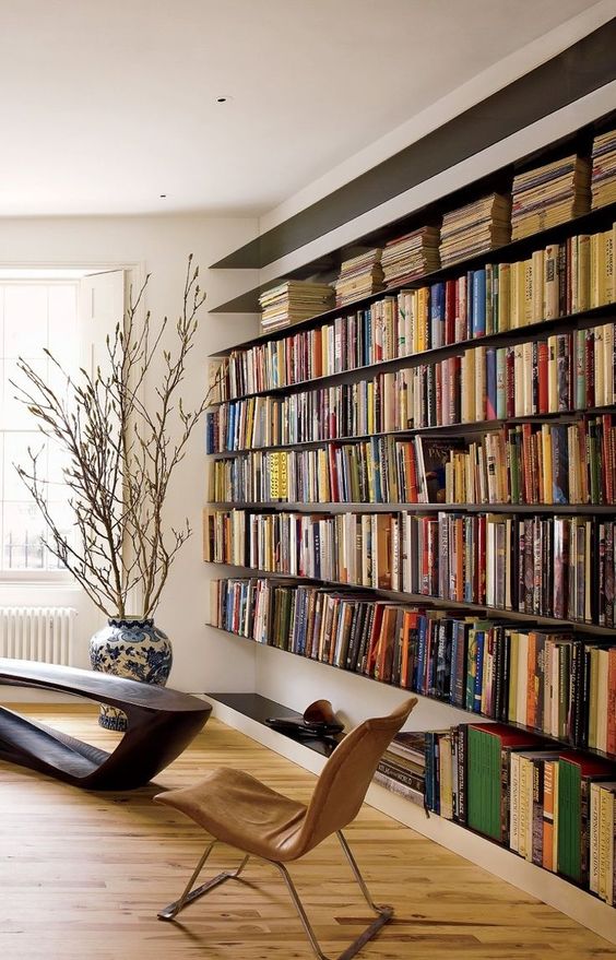 a modern refined home library with an oversized floating bookshelf unit, a dark wooden table and a leather chair