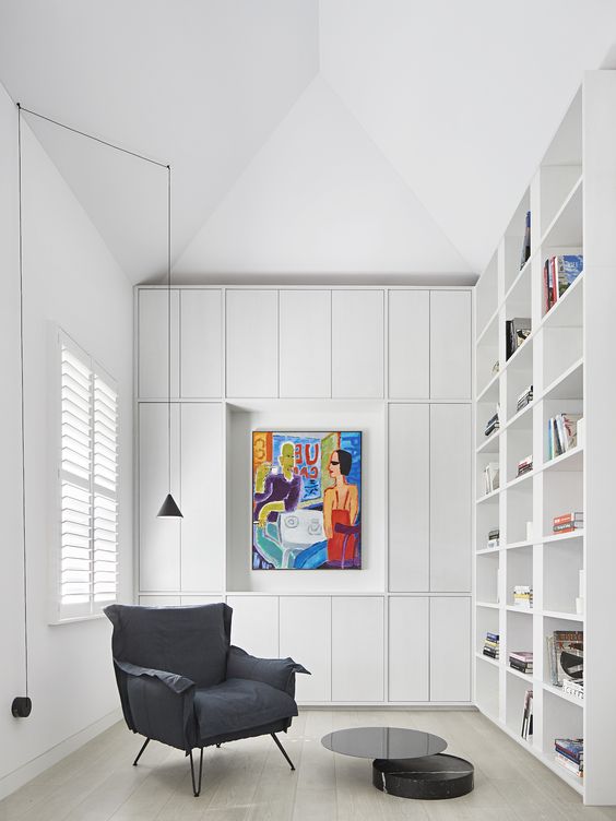 A modern white home library with built in bookshelves, a grey chair and some closed up storage units