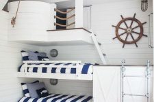 a nautical kids’ bedroom clad with white shiplap, with built-in bunk beds, a metal ladder and a ship part on top