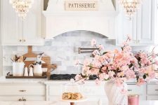 a romantic glam kitchen with white vintage cabinetry, white marble tiles, stools with gold rings, crystal chandeleirs and pink blooms