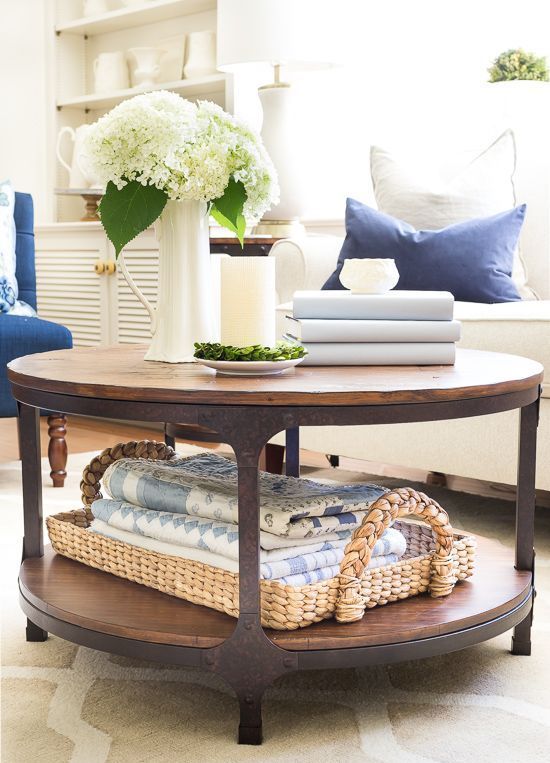 a rustic coffee table with a woven tray with towels, a stack of books, a vase with white blooms, a candle and greenery