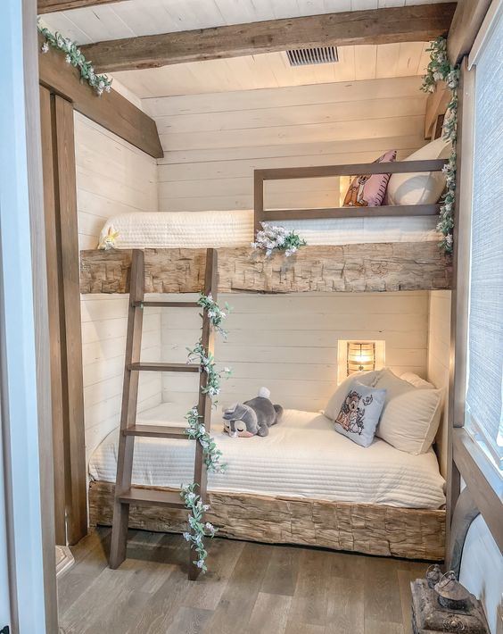 A rustic kids' bedroom with built in rough wood beds, a ladder covered with greenery and blooms, built in sconces and beams on the ceiling