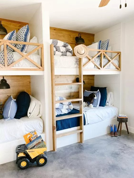 A rustic kids' room with four built in bunk beds, white, blue and navy bedding, a ladder and lots of pillows and black sconces
