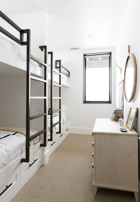 A small neutral kids' bedroom with built in bunk beds, black ladders, a large white dresser and a round mirror