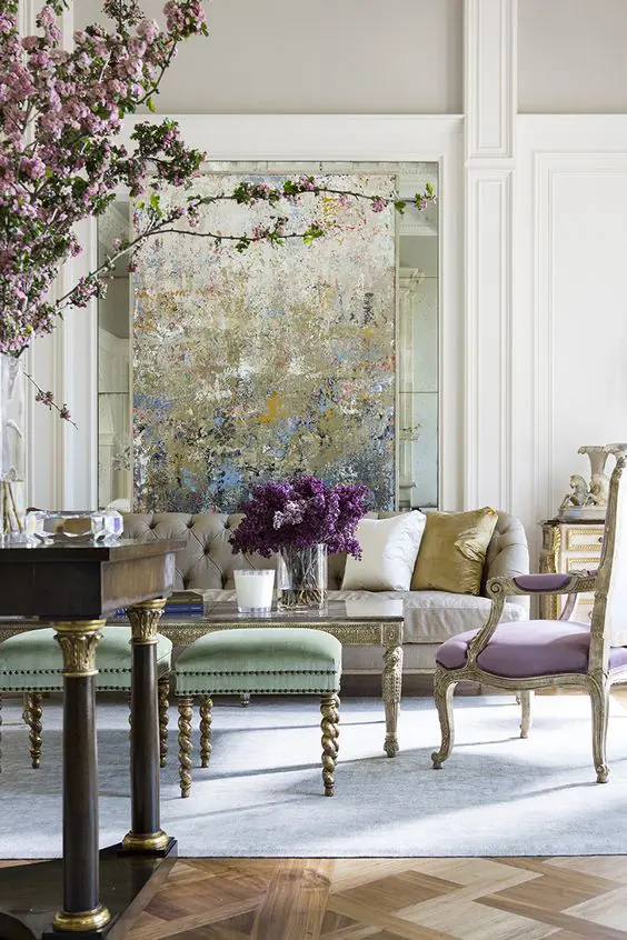 A sophisticated pastel space with an impressionalist artwork, a neutral sofa, an antique lilac chair, green stools and a dark stained table