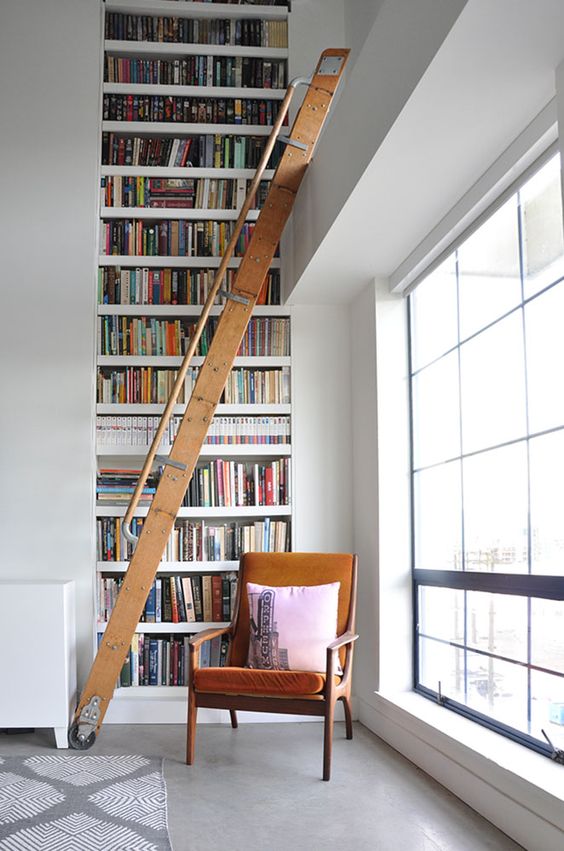 A stylish modern library nook with a double height bookcase, a staircase and a comfy chair in rust