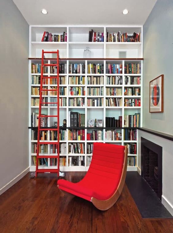 A stylish modern library with a large built in bookcase unit, a built in fireplace and a red curved chair and a red staircase