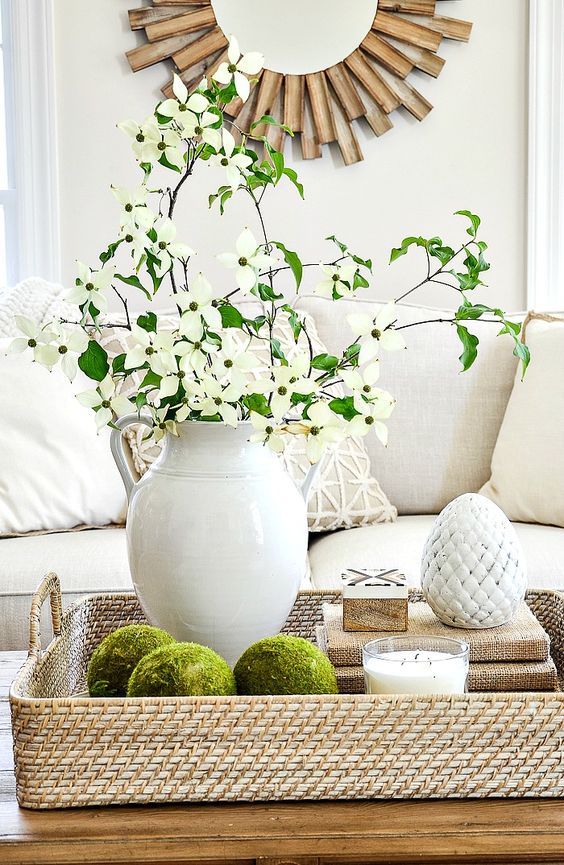 a woven tray with moss balls, a stack of books, a candle, a porcelain egg and a white jug with fresh blooms and greenery