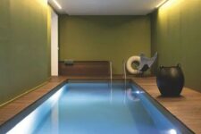 an indoor pool space with green walls, built-in lights, a pool and a wooden deck, a chair and a large vase is a bold idea for a modern home