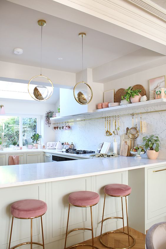An off white glam kitchen with white marble countertops and a backsplash, unique pendant sphere lamps, pink stools