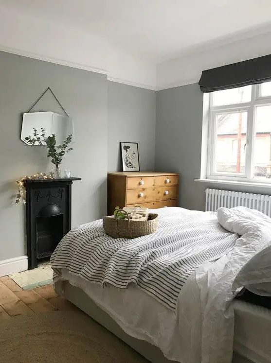 A Nordic bedroom with grey walls, a vintage built in hearth, a bed with neutral and printed bedding, a stained dresser and a mirror