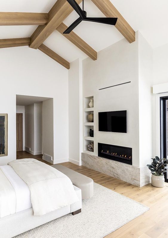 A beautiful and sleek creamy bedroom with wooden beams, a built in fireplace, built in shelves and potted plants
