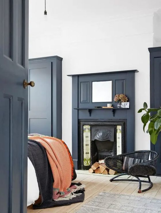 a chic bedroom done with touches of navy, a built-in fireplace, a bed with bright bedding and a black rattan chair plus a pendant bulb