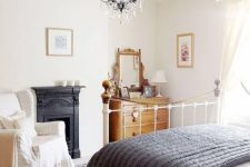 a chic vintage bedroom with a metal built-in fireplace, a white metal bed, a stained dresser, a vintage chair and a beautiful crystal chandelier