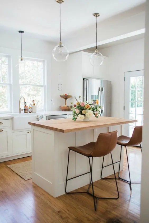 a classy and clean white kitchen with shaker style lower cabinets, a small kitchen island with a stained countertop and leather stools