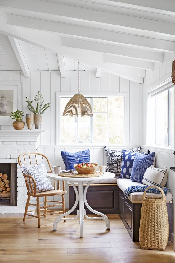 a coastal space with a white brick clad fireplace, a banquette corner seating, a round table and a wooden chair is welcoming