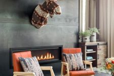 a contemporary fireplace nook with a concrete clad fireplace, a faux taxidermy piece, orange chairs and pillows plus cowboy boots