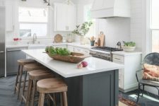 a farmhouse kitchen with white cabinets and a large hood, pendant lamps, a graphite grey kitchen island and wooden stools