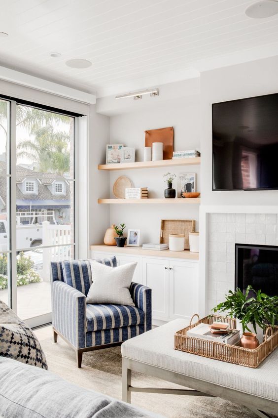 A farmhouse space wiht a white brick clad fireplace, built in shelves and cabinets, a striped chair and a large ottoman and greenery