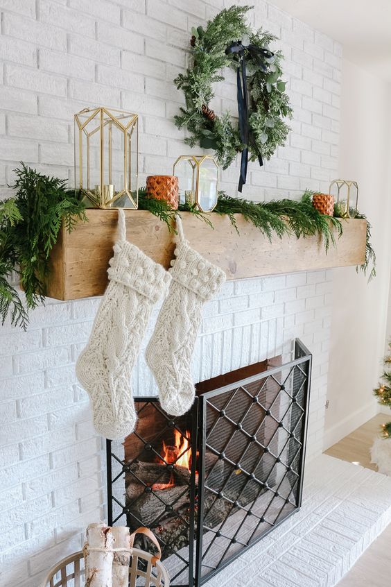 a fireplace styled with evergreens, an evergrene wreath and candle lanterns plus stockings for a cool look