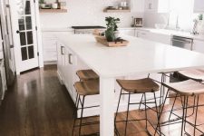a large airy white kitchen with shaker style cabinets, stained shelves and a hood, a large kitchen island with a storage cabinet and an eating space