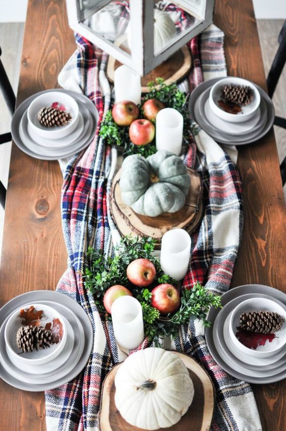 a lovely fall or Thanksgiving tablescape with a plaid runner, greenery, apples, pillar candles, neutral pumpkins and pinecones in a plate