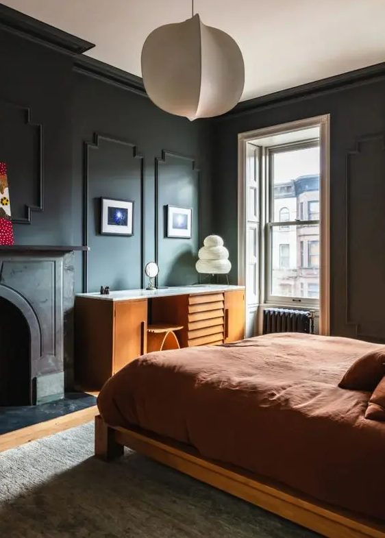 A lovely moody bedroom with black trim walls, a non working fireplace, a stained bed with rust bedding, a stained desk