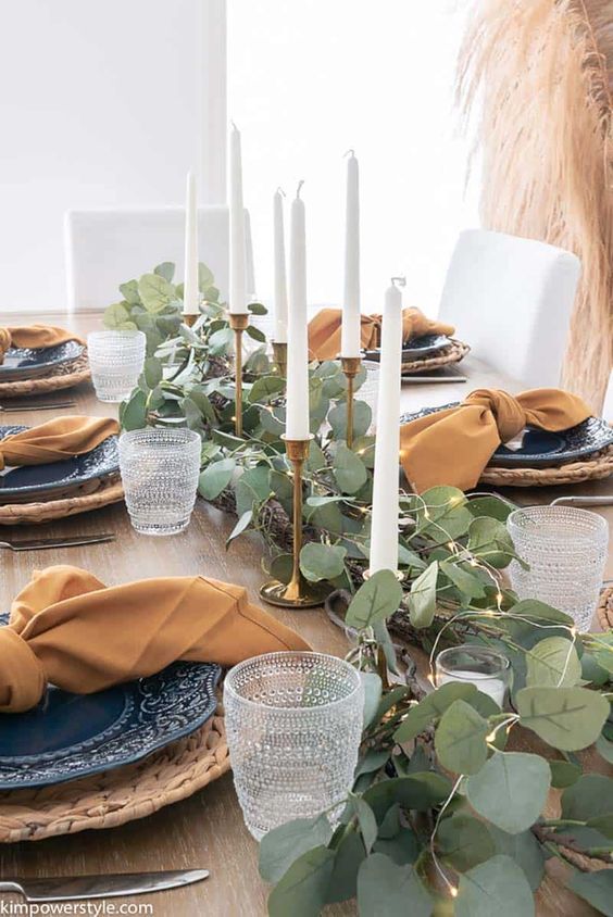 a modern farmhouse Thanksgiving tablescape with a greenery and light runner, candles, blue plates, woven chargers and terracotta napkins
