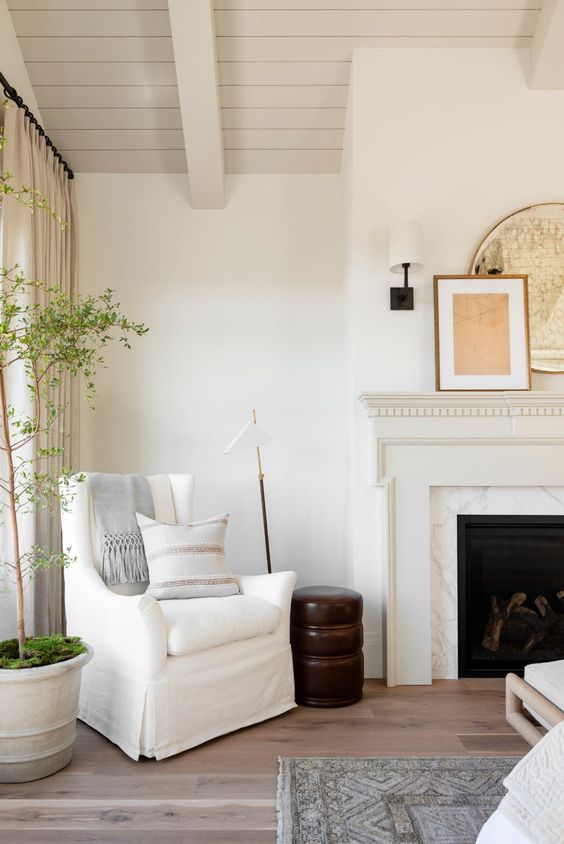 a refined space with a marble clad fireplace, a neutral wooden mantel, a white chair with a pillow and a wooden side table