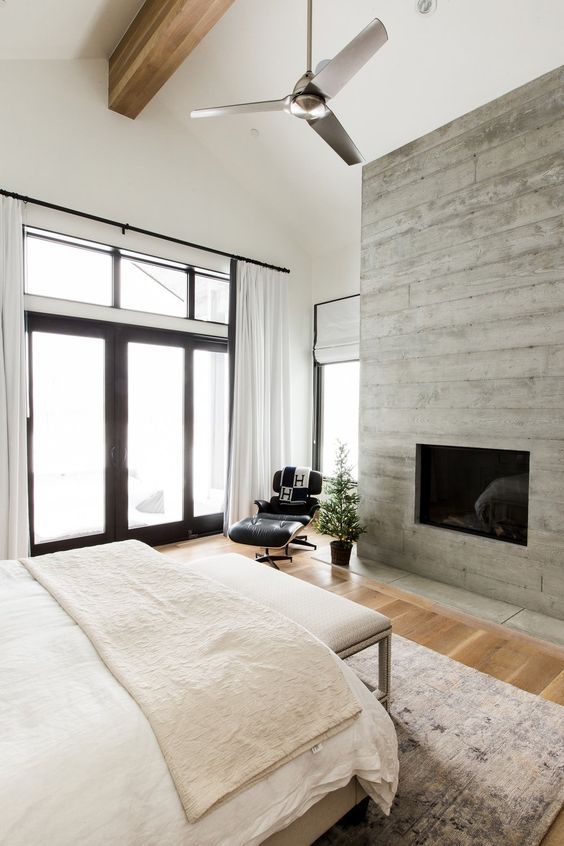 a serene bedroom with a large window, a fireplace clad with wood, a bed with neutral bedding and a bench