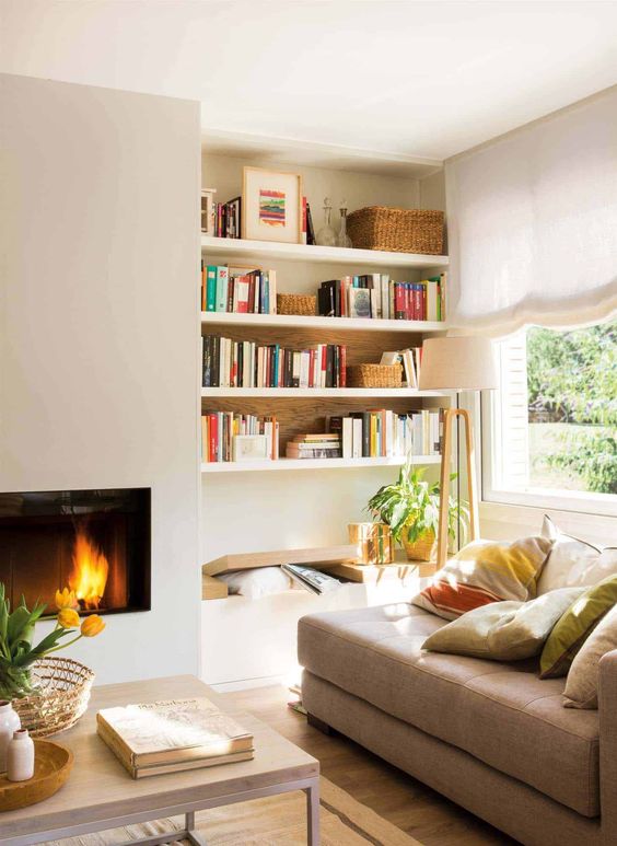 A sleek built in fireplace, a bench and built in shelves, a tan couhc with pillows form a cozy and welcoming reading nook