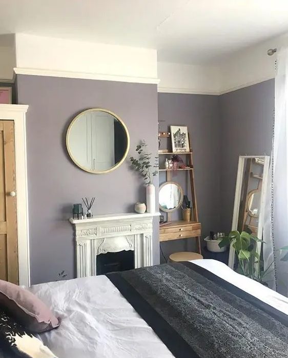 A small yet cozy bedroom with lavender walls, a refined built in fireplace, a mini stained wood shelving unit and a round mirror