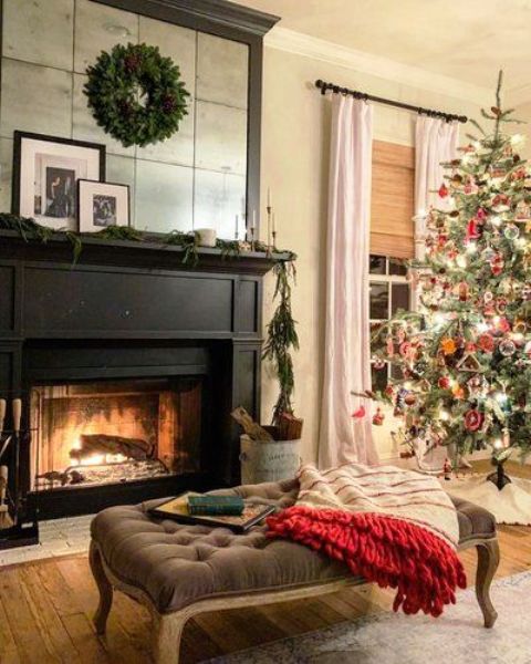 a stylish and welcoming fireplace nook with a black fireplace, a mirror over it, a vintage ottoman and a blanket plus a greenery wreath