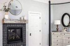 a vintage farmhouse bedroom with a black metal canopy bed, a built-in fireplace with black tiles, a round mirror, a white dresser and a wooden chandelier
