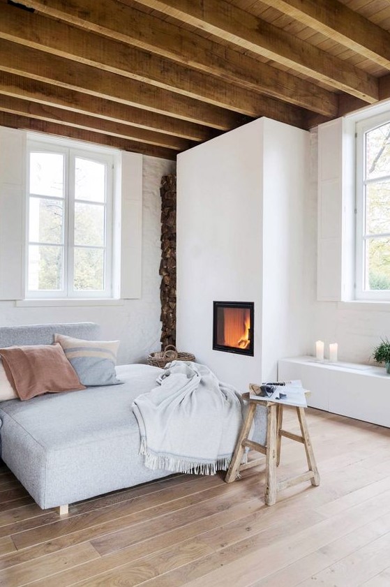 A welcoming and neutral looking bedroom with a built in fireplace, a bed, a stool, a storage unit with candles and wooden beams on the ceiling