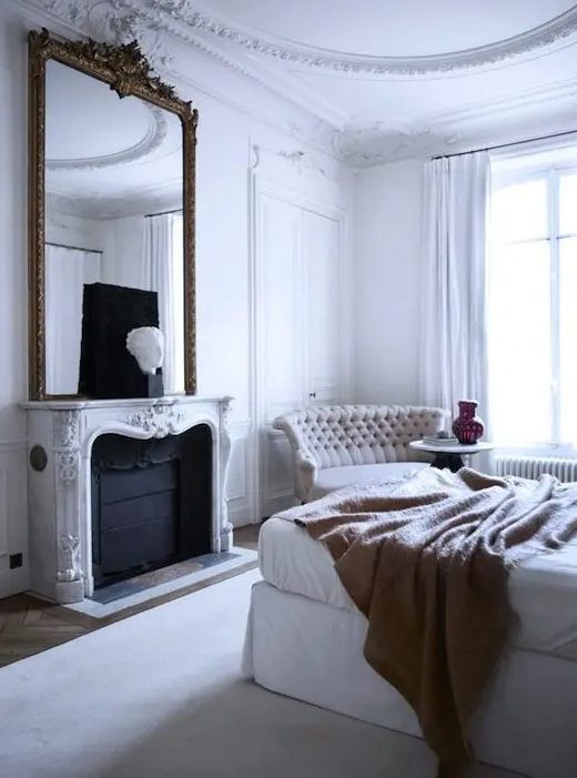 a white Parisian bedroom with a statement mirror over the fireplace, an upholstered bed, a chic tufted sofa and much natural light