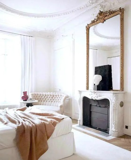 An exquisite bedroom with a French feel, with a non working fireplace, a white bed with a tan blanket, a refined loveseat and an oversized mirror