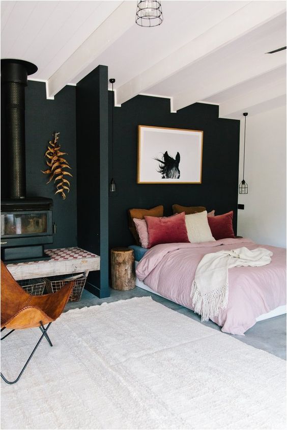 An eye catchy bedroom with a black accent wall, a black hearth and a bed with pink and burgundy bedding, an amber chair
